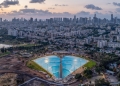 צילום אווירי של בריכת הגלים המלאכותית בפארק דרום בתל אביב. במרכז התמונה בריכה בצורת מניפה, מוקפת בשטחי עפר ופיתוח של אזור שכונת הארגזים, כשברקע נראה קו הרקיע של מגדלי גוש דן.