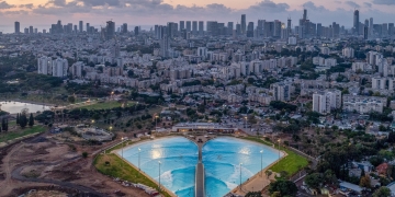 צילום אווירי של בריכת הגלים המלאכותית בפארק דרום בתל אביב. במרכז התמונה בריכה בצורת מניפה, מוקפת בשטחי עפר ופיתוח של אזור שכונת הארגזים, כשברקע נראה קו הרקיע של מגדלי גוש דן.