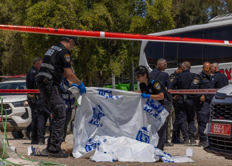 שוטרי משטרת ישראל ואנשי זיהוי פלילי במדים כהים עומדים בזירת אירוע מגודרת בסרט חוסם אדום-לבן. שוטר ושוטרת עוטי כפפות מכחילות פורסים יריעת פלסטיק לבנה עם הדפס כחול מעל גופה על הקרקע, שרק כף רגל חשופה מבצבצת ממנה. סביבם ניידות משטרה לבנות, אוטובוס ועצים ירוקים ברקע, ותוויות מספור צהובות של ראיות מפוזרות על הקרקע.